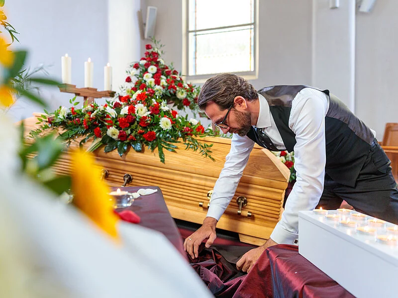 Bestatter dekoriert in der Trauerhalle Blumen, Kerzen, Sarg und Samt für die Aufbahrung.