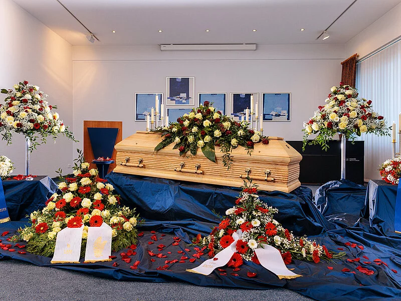 Geschlossene Aufbahrung eines Sargs in einer Trauerhalle mit Blumenschmuck und blauem Samt.