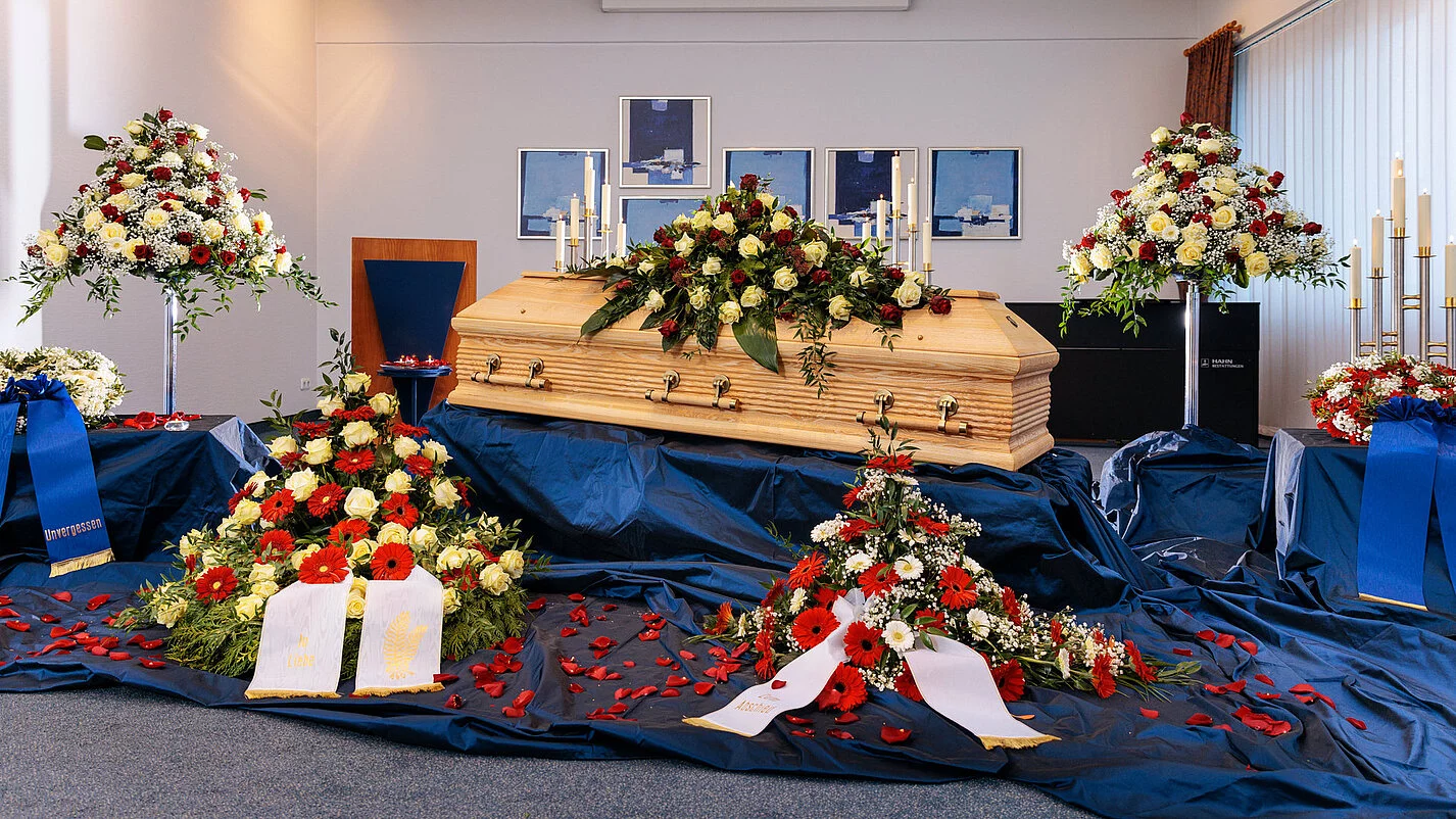Geschlossene Aufbahrung eines Sargs in einer Trauerhalle mit Blumenschmuck und blauem Samt.