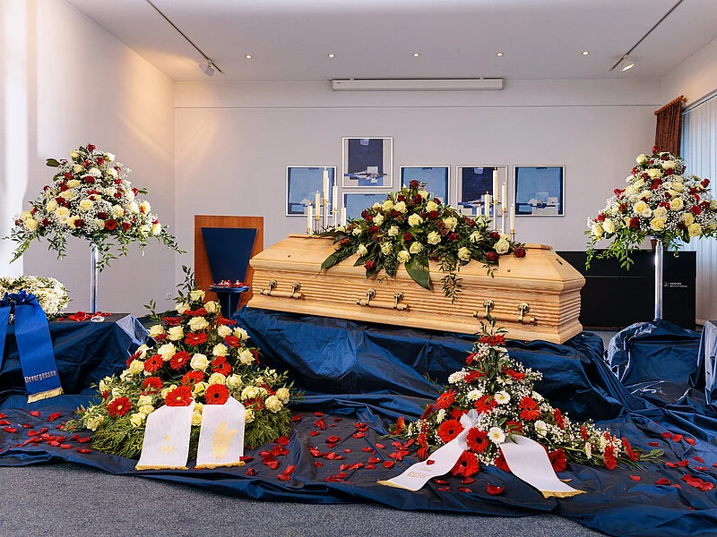 Geschlossene Aufbahrung eines Sargs in einer Trauerhalle mit Blumenschmuck und blauem Samt.