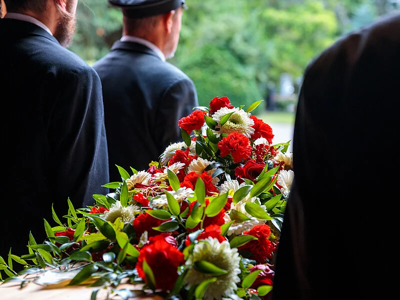 Vier Sargträger in Schwarz tragen einen Holzsarg mit aufliegendem Blumenschmuck