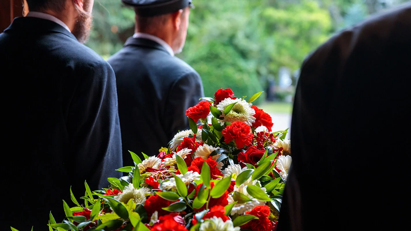 Vier Sargträger in Schwarz tragen einen Holzsarg mit aufliegendem Blumenschmuck