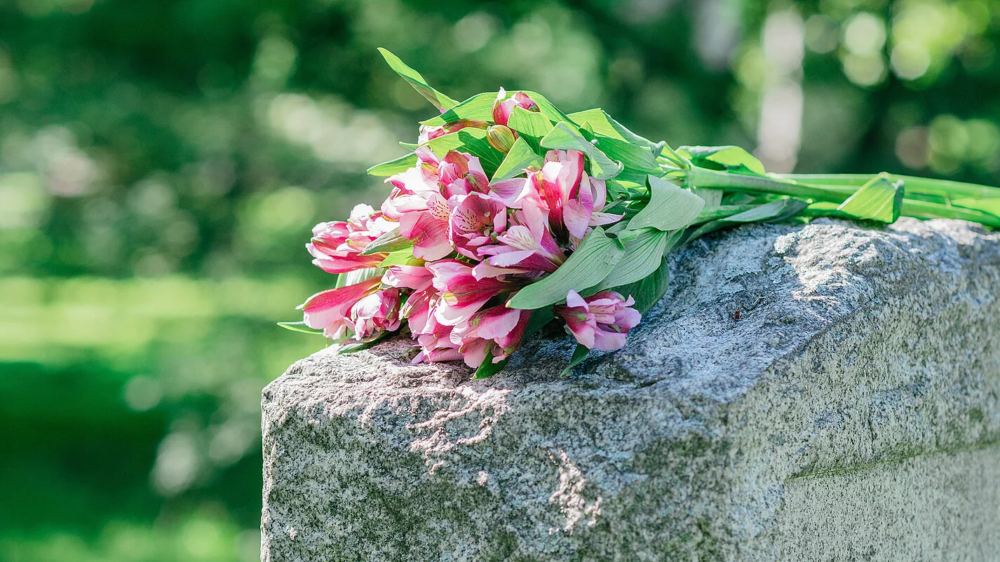 Grabstein auf dem ein Straus Blumen mit pinken Blüten liegt.