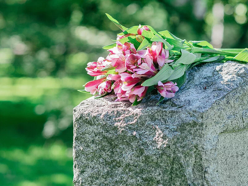 Grabstein auf dem ein Straus Blumen mit pinken Blüten liegt.