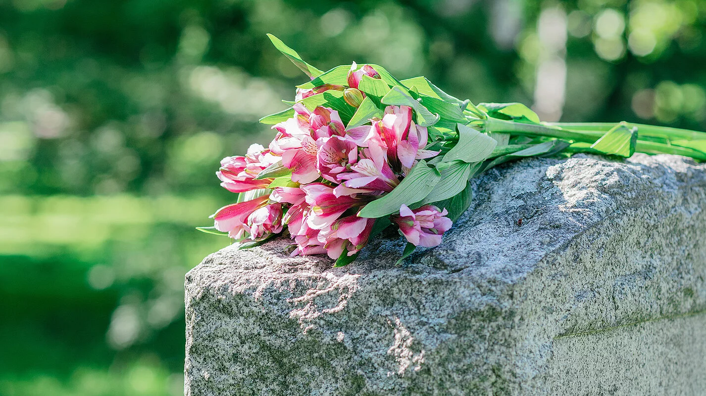 Grabstein auf dem ein Straus Blumen mit pinken Blüten liegt.