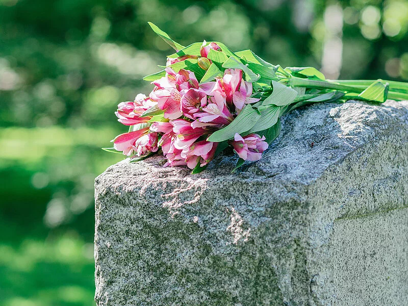 Grabstein auf dem ein Straus Blumen mit pinken Blüten liegt.