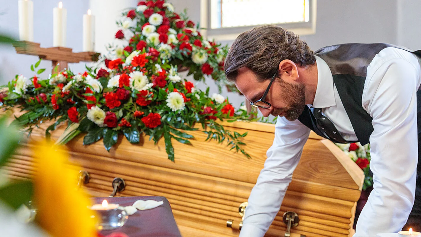 Bestatter dekoriert in der Trauerhalle Blumen, Kerzen, Sarg und Samt für die Aufbahrung.