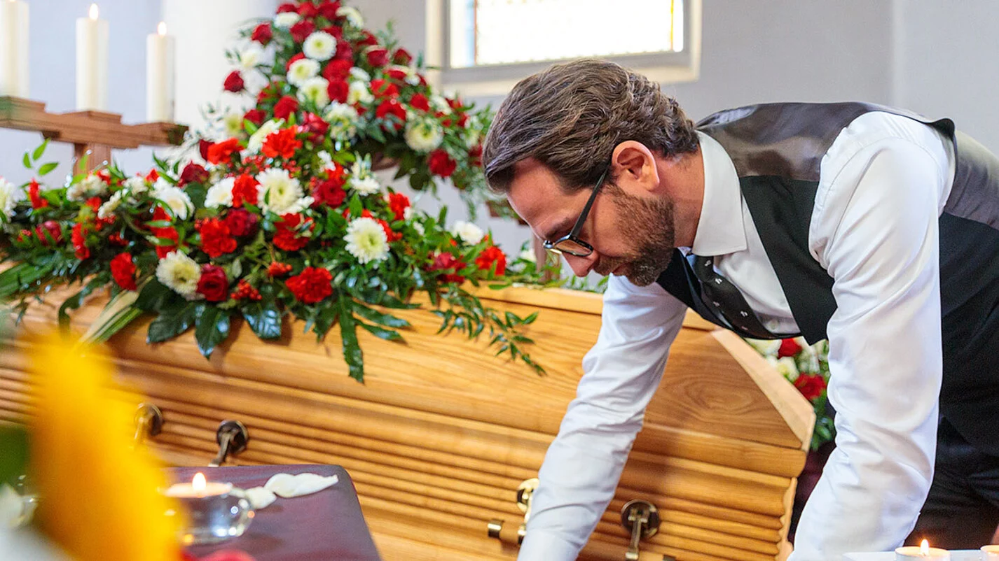 Bestatter dekoriert in der Trauerhalle Blumen, Kerzen, Sarg und Samt für die Aufbahrung.