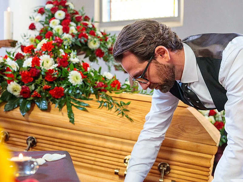 Bestatter dekoriert in der Trauerhalle Blumen, Kerzen, Sarg und Samt für die Aufbahrung.
