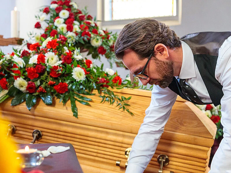 Bestatter dekoriert in der Trauerhalle Blumen, Kerzen, Sarg und Samt für die Aufbahrung.