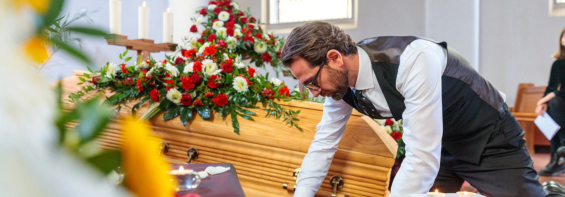 Bestatter dekoriert in der Trauerhalle Blumen, Kerzen, Sarg und Samt für die Aufbahrung.