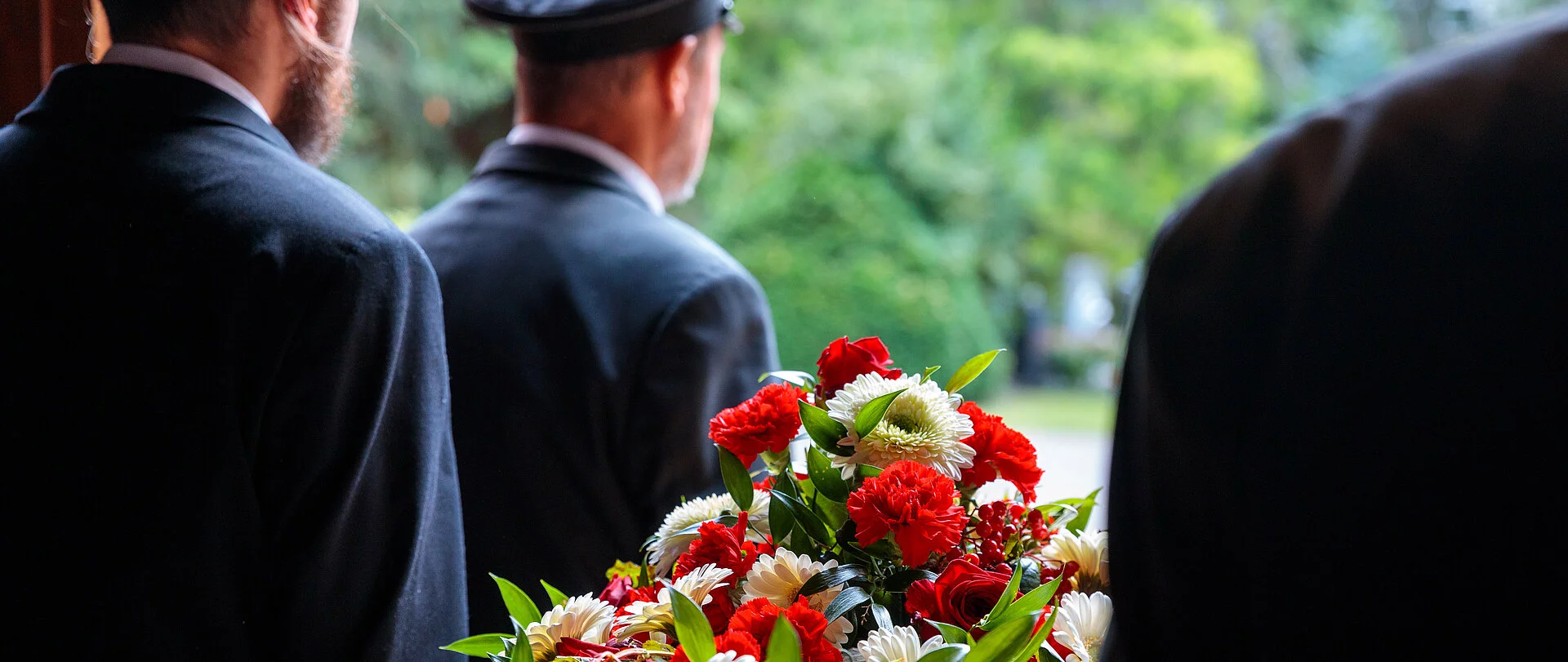 Vier Bestatter tragen einen Holzsarg mit aufgelegtem Blumenschmuck.